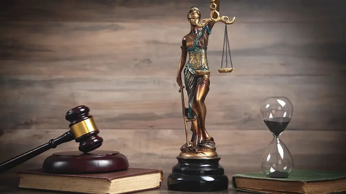 Temida trzyma symbole prawa. Figurka jest na biurku, na nim znajdują się kodeksy prawa z klepsydrą.